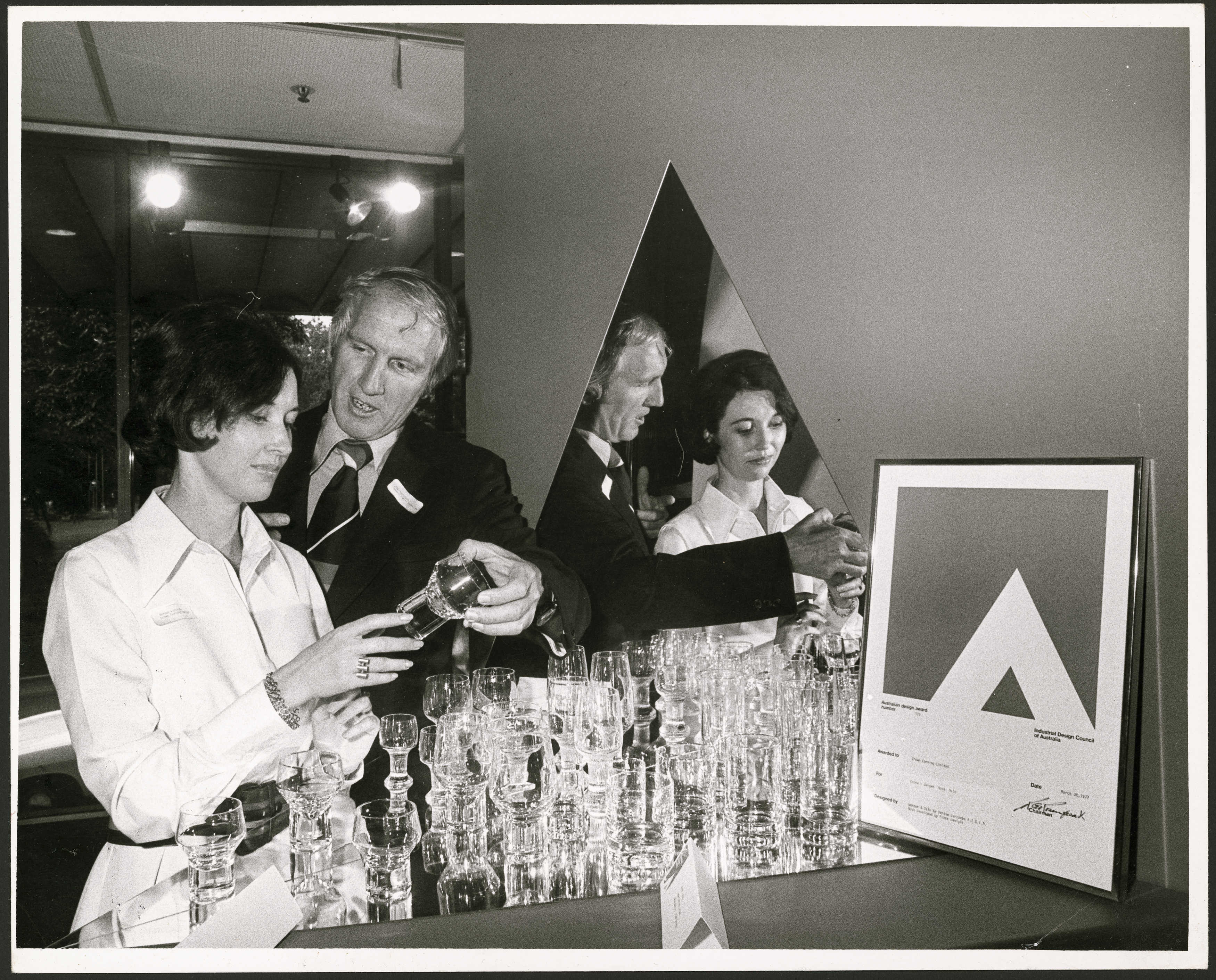 Black and white photograph of a woman and man, both holding a drinking glass. There is a large display of drinking glasses in front of them and a framed award on the right of the image.